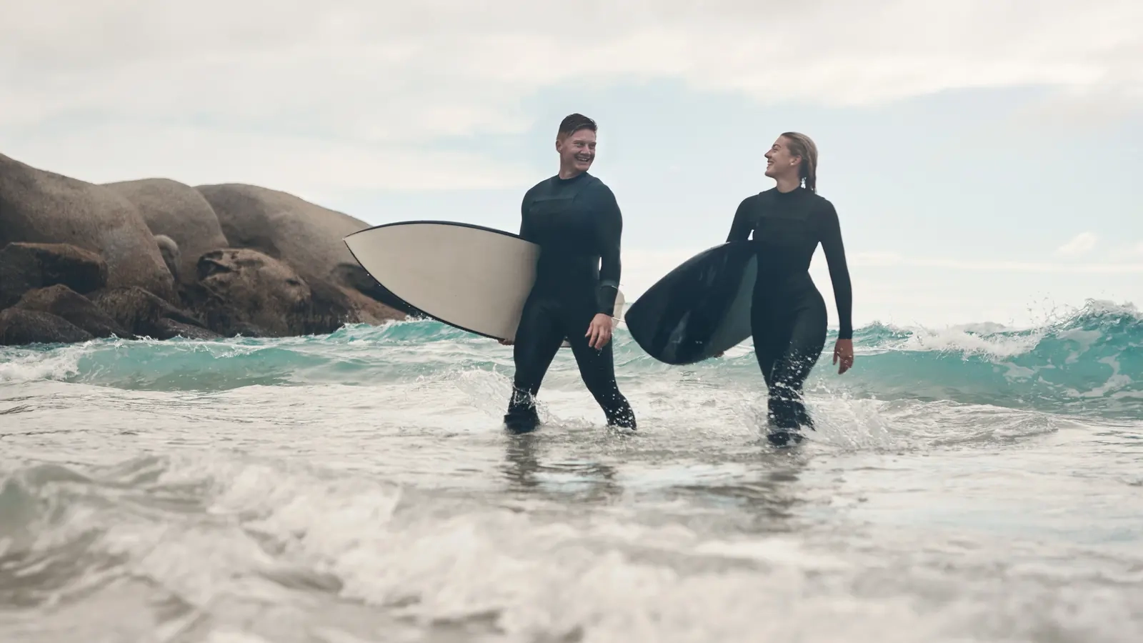 A smiling couple in wetsuits wading out into the surf carrying their surfboards
