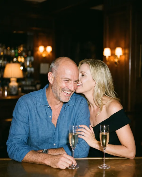 A blonde woman whispering to a laughing older man at a bar