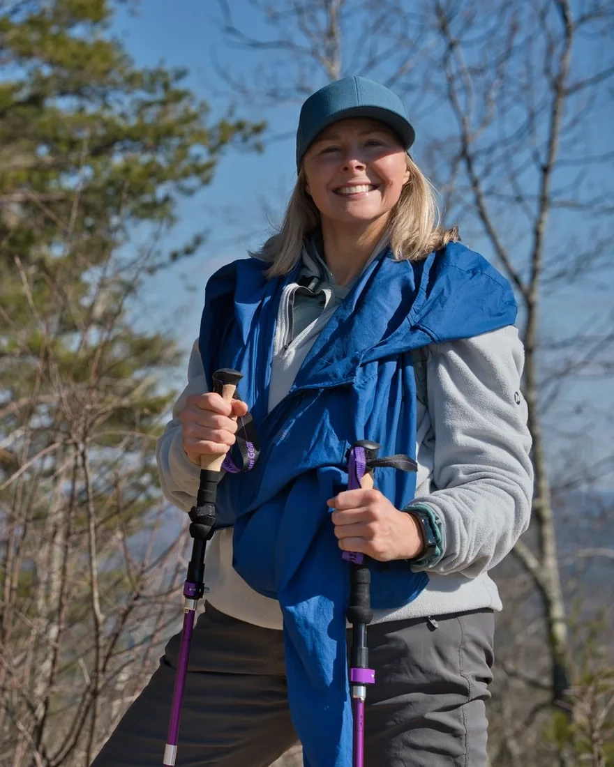 A smiling woman in a cap and blue jacket holding trekking poles on a sunny mountain trail