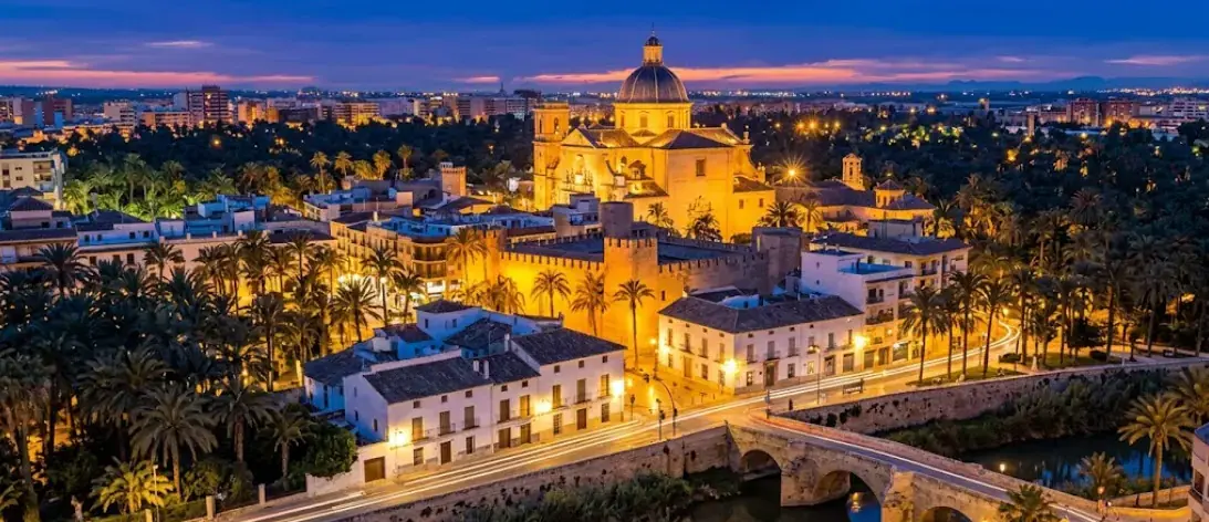 Vista aérea de Elche al anochecer, con la basílica de Santa María iluminada rodeada del palmeral