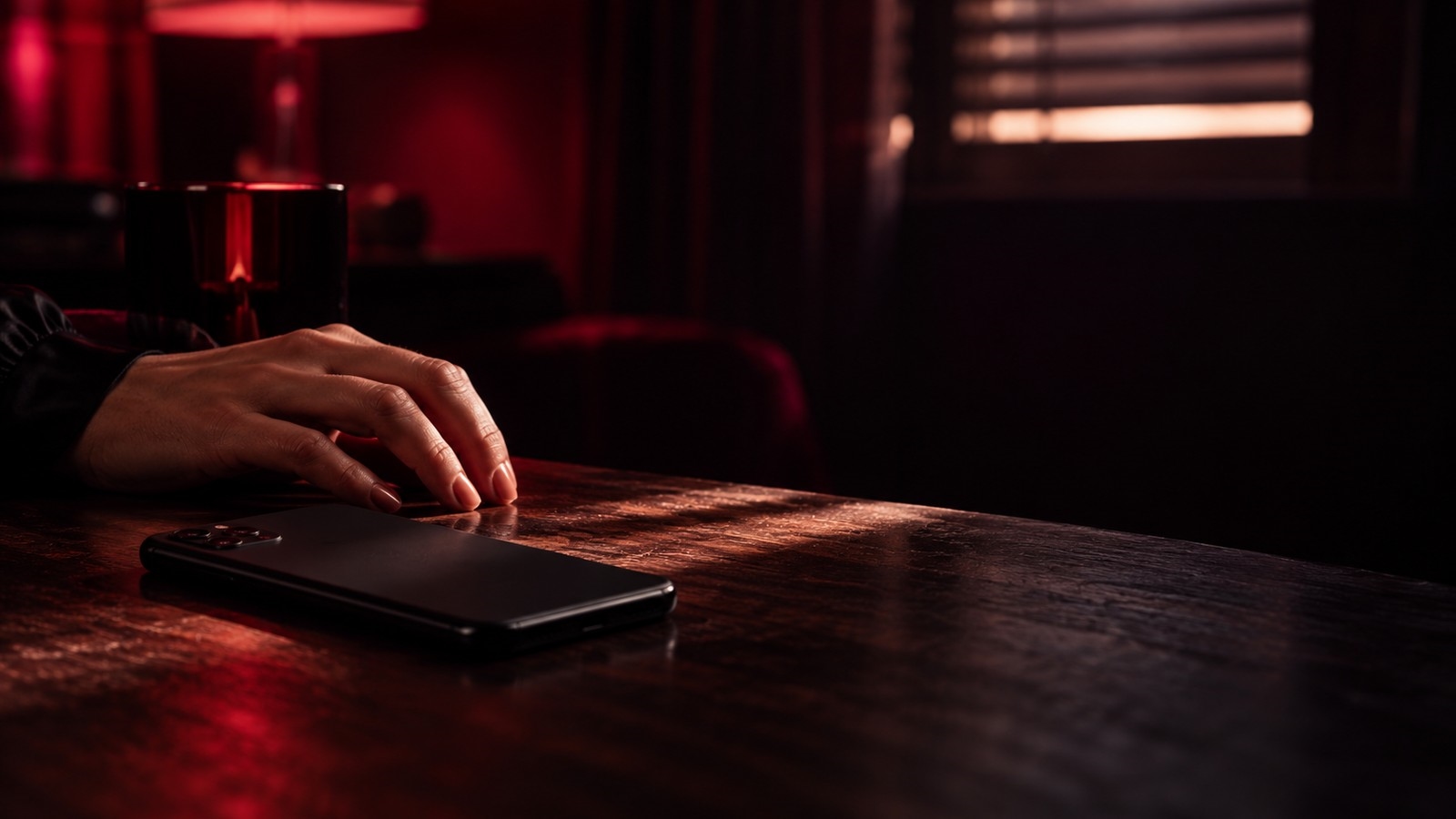 Una mano reposa junto a un teléfono móvil colocado bocabajo sobre una mesa de madera, en una habitación en penumbra con luz roja y una persiana entornada al fondo