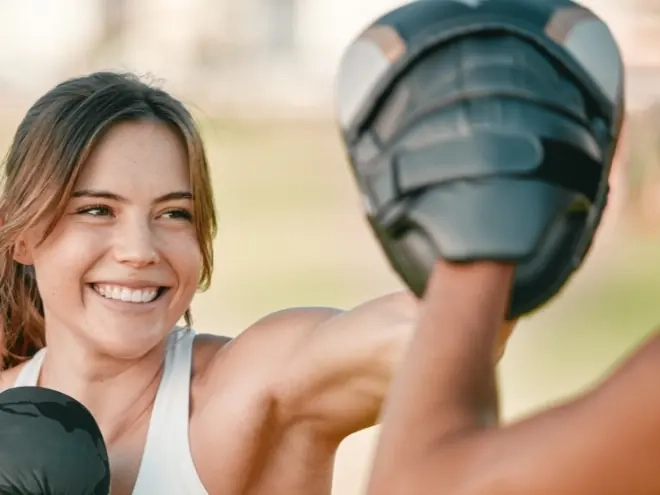A smiling woman punching focus pads held by her trainer