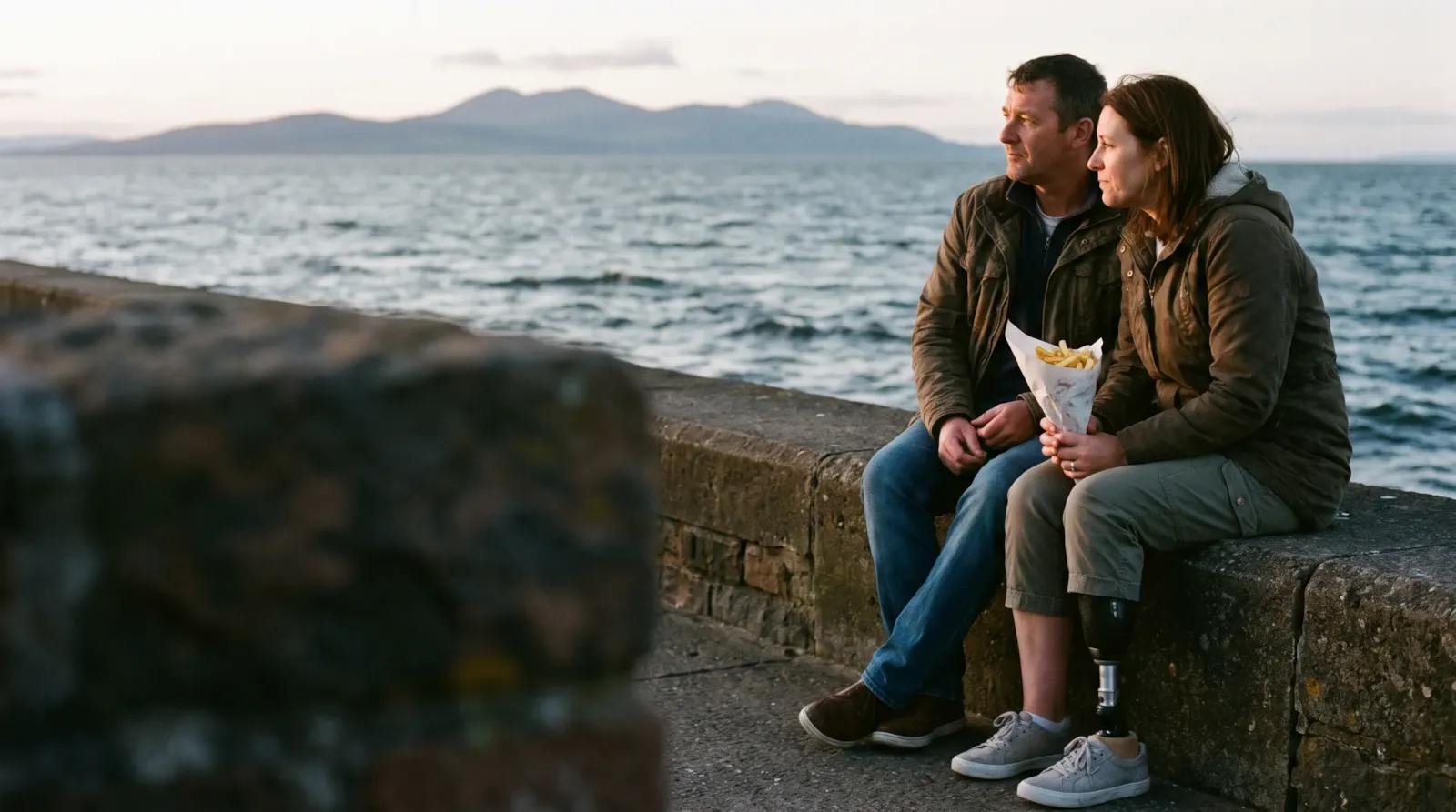 A couple sharing chips on the sea wall at Ayr at sunset, the Isle of Arran behind them; the woman has a prosthetic leg