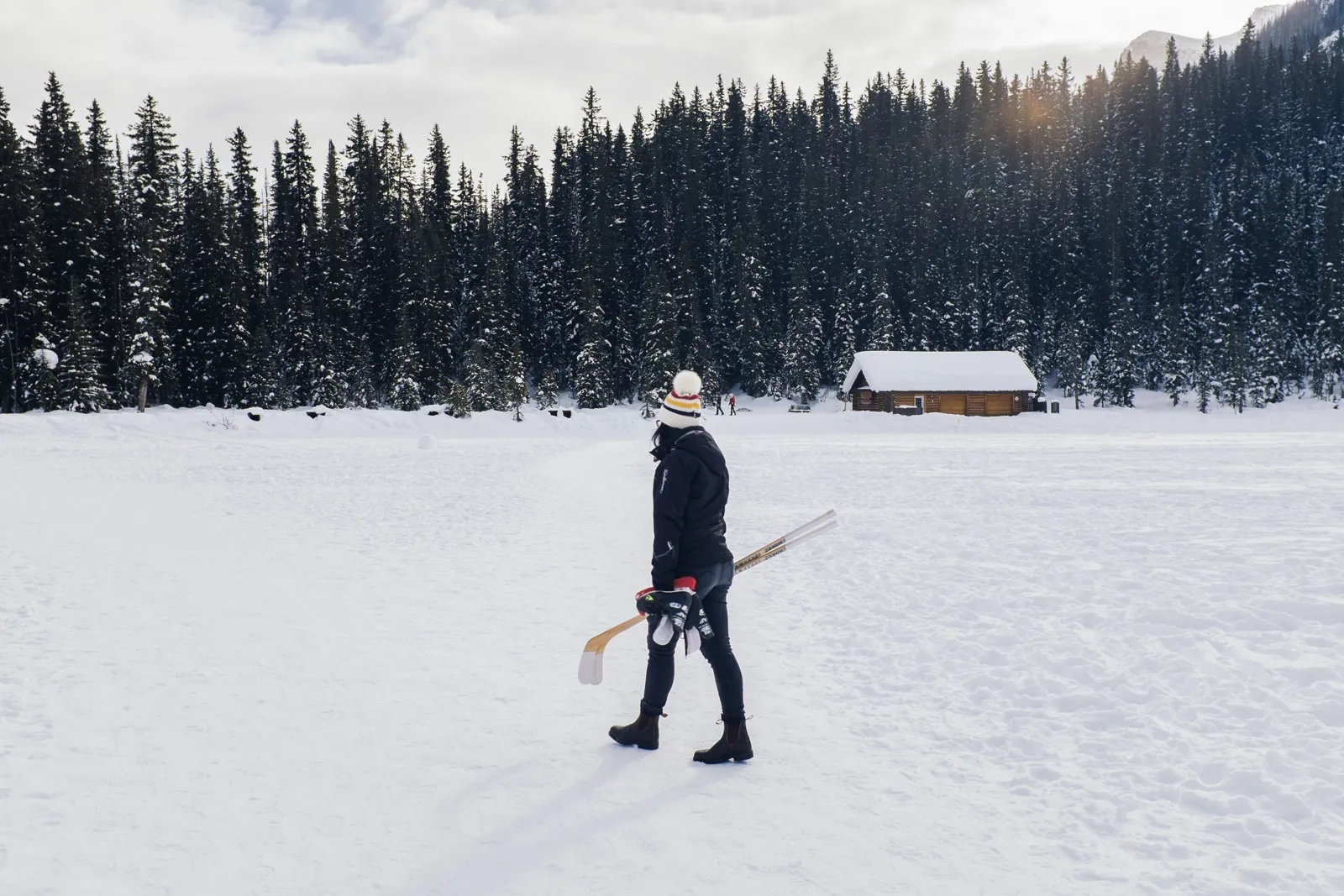 Person holding a hockey stick on a snowy outdoor rink with a forest and cabin behind