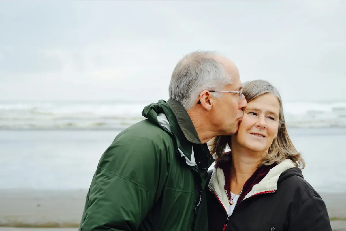 A man in a green jacket kissing a smiling woman on the cheek by the sea