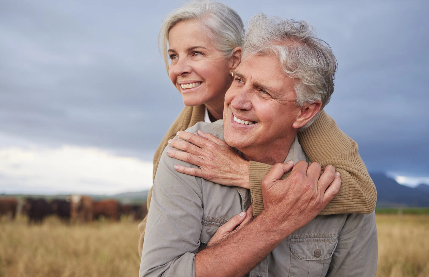 A smiling gray-haired woman with her arms around a man's shoulders in a field under a stormy sky