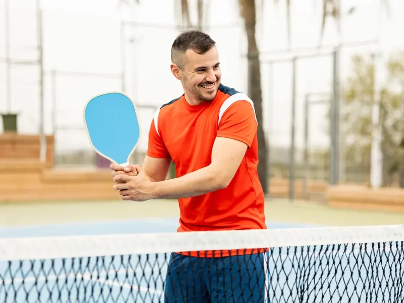 A smiling man holding a blue pickleball paddle at the net