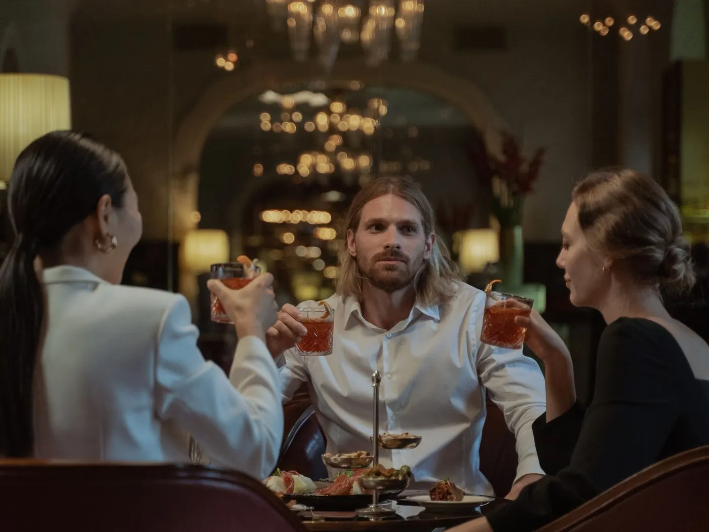 Three friends in a grand lounge bar raising cocktails to each other: a woman in a white jacket, a long-haired man in a white shirt and a woman in black