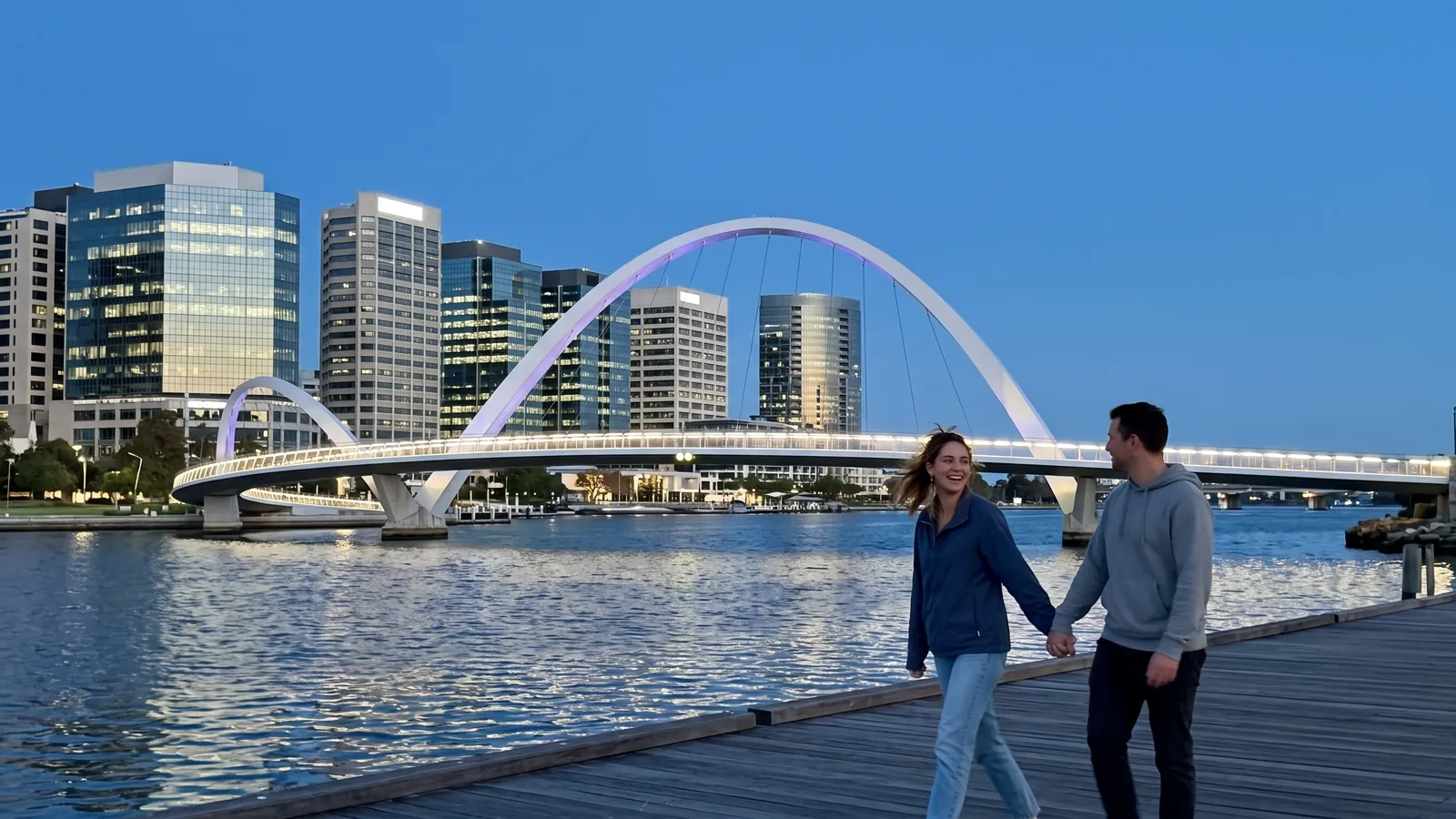 A couple walks hand in hand along the Elizabeth Quay promenade in Perth at dusk, the curved footbridge and the lit city towers behind them (AI-generated image)