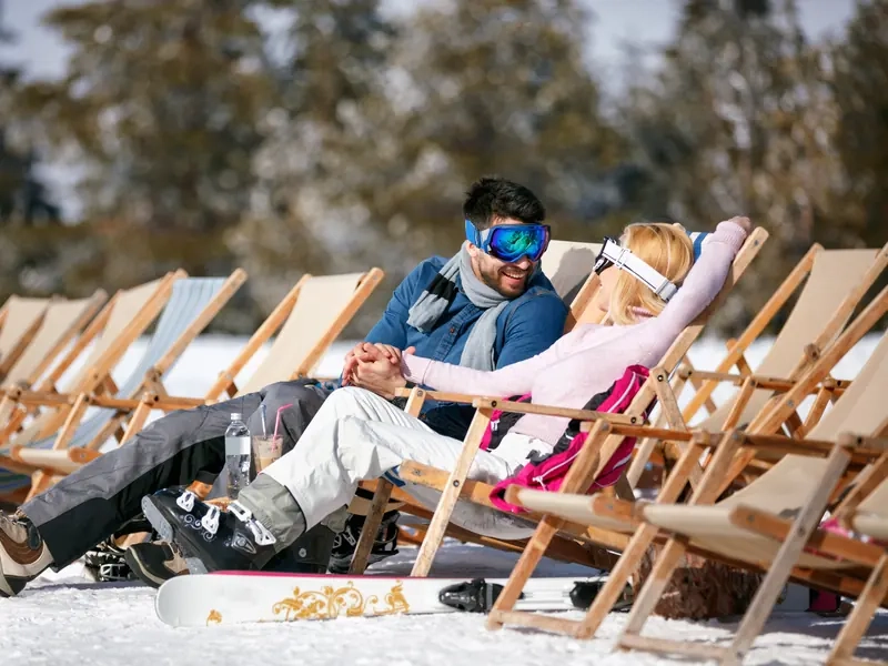 A couple in ski gear holding hands and relaxing in deckchairs in the snow