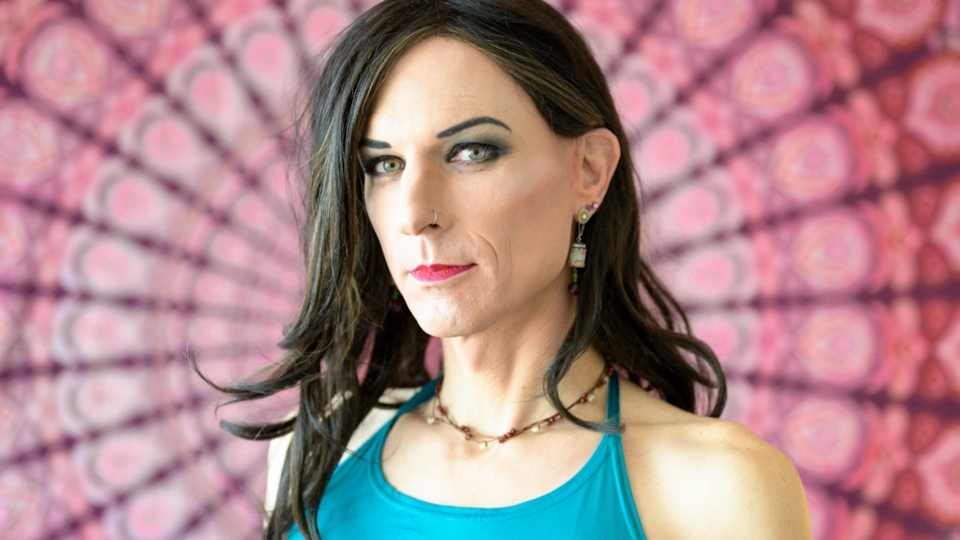 A trans woman with long dark hair and a turquoise top, looking straight at the camera in front of a pink mandala