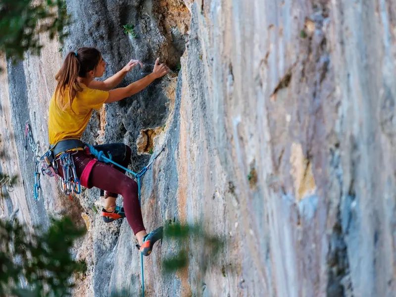 A woman in a yellow T-shirt lead climbing a tall limestone wall, reaching for the next hold