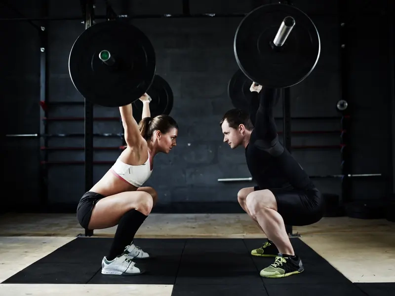 A man and woman holding barbells overhead in matching squats, face to face