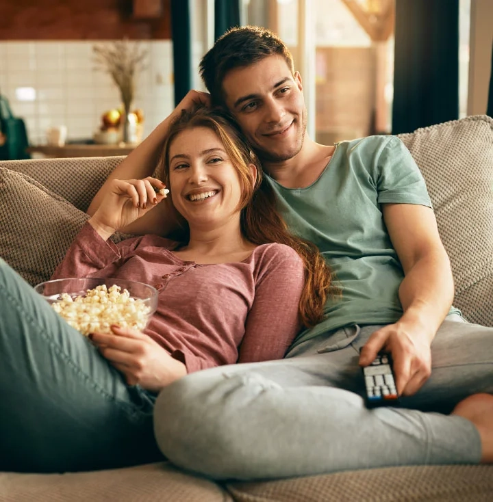 A young couple relaxing on a sofa with a bowl of popcorn and the TV remote, both smiling