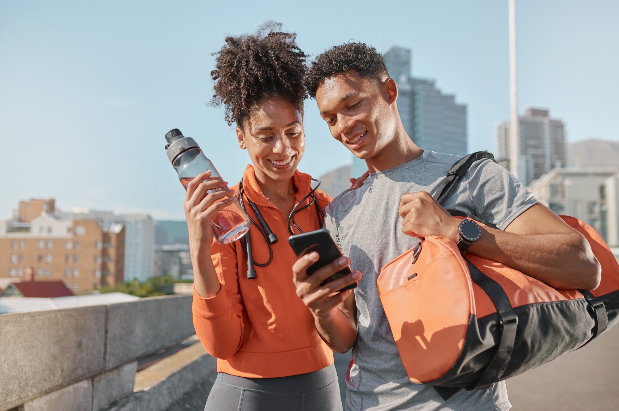 A smiling woman and man in workout clothes looking at a phone together on a sunny city rooftop