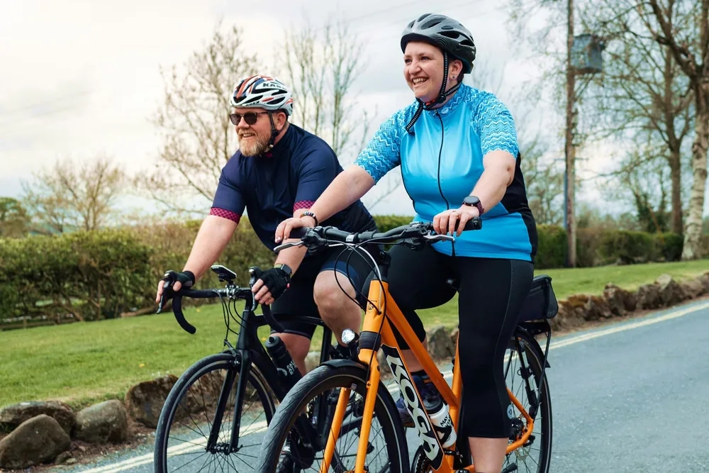 A man and a woman in cycling helmets riding side by side along a sunny country lane