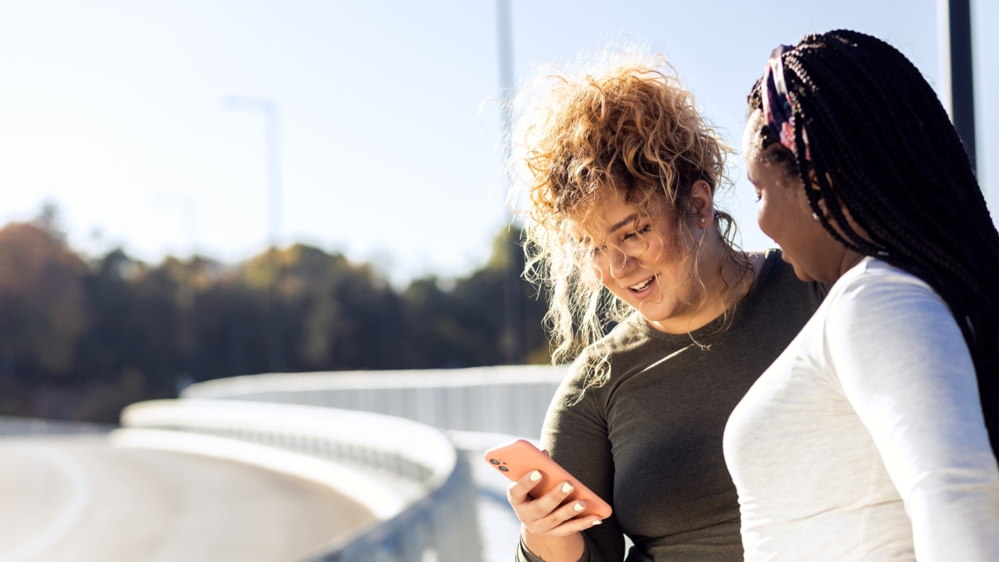 Two friends smiling and looking at a phone together outdoors