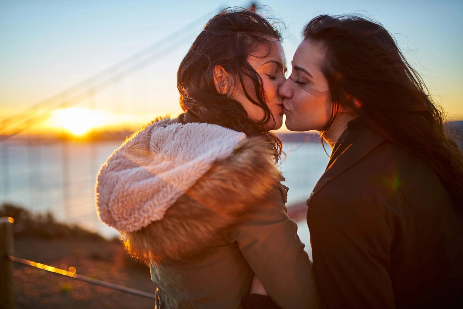Two women kissing at sunset by the water, the sun low behind them