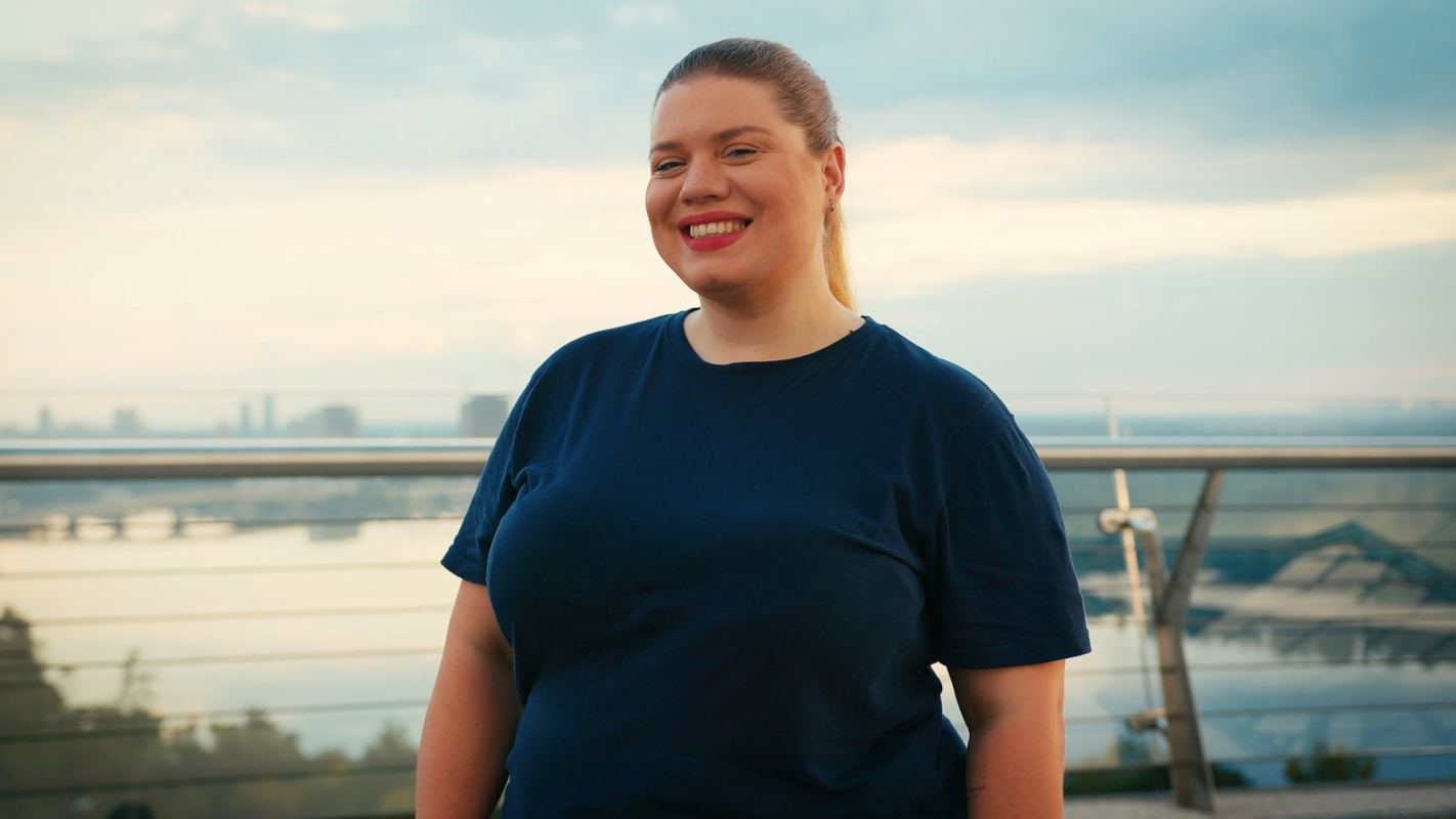 A woman smiling confidently outdoors with a city waterfront view behind her