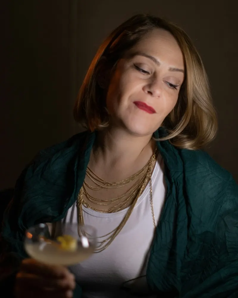 A woman in a green jacket and gold necklaces smiling with her eyes closed, cocktail in hand, in a dimly lit bar