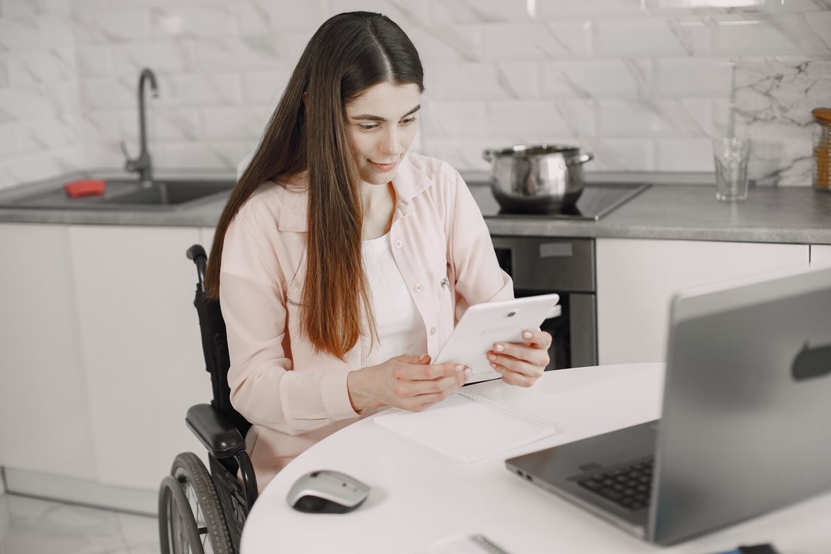 A woman in a wheelchair smiling while looking at a tablet in a bright kitchen