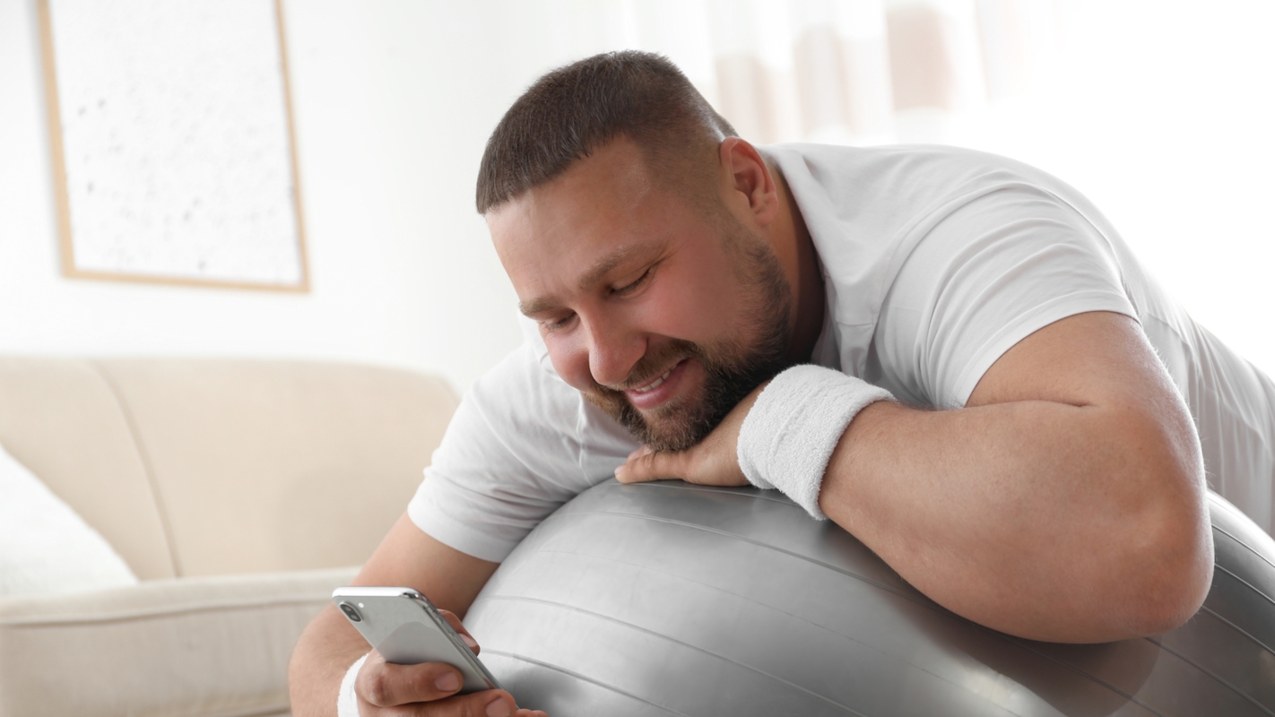 A man smiling at his phone while leaning on an exercise ball at home