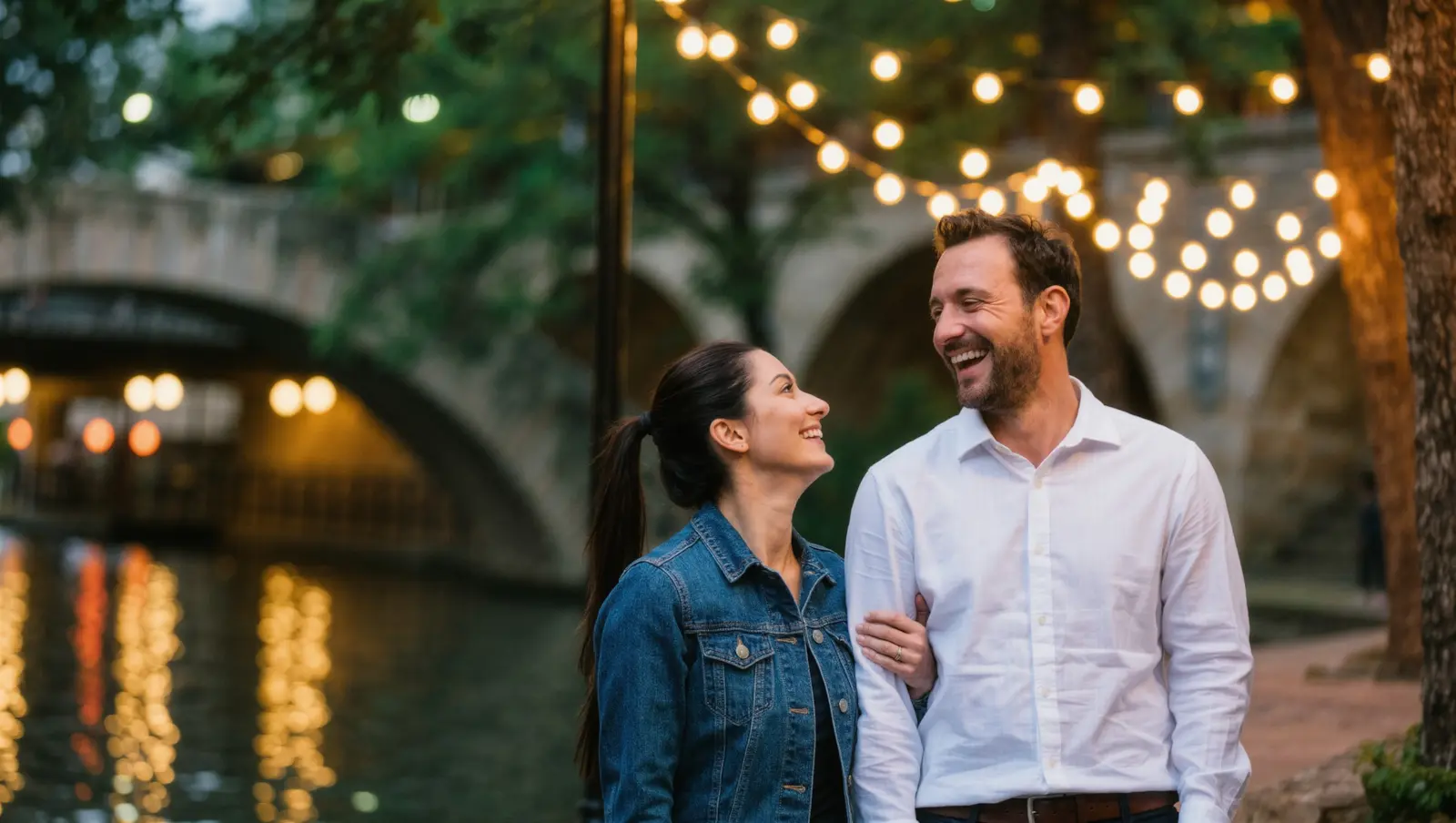 A couple laughing together arm in arm on a lantern-lit riverside walk with a stone bridge behind them