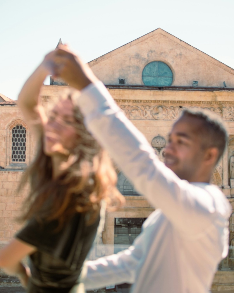Una pareja baila frente a una iglesia de piedra; él, de camisa blanca, le da una vuelta a ella