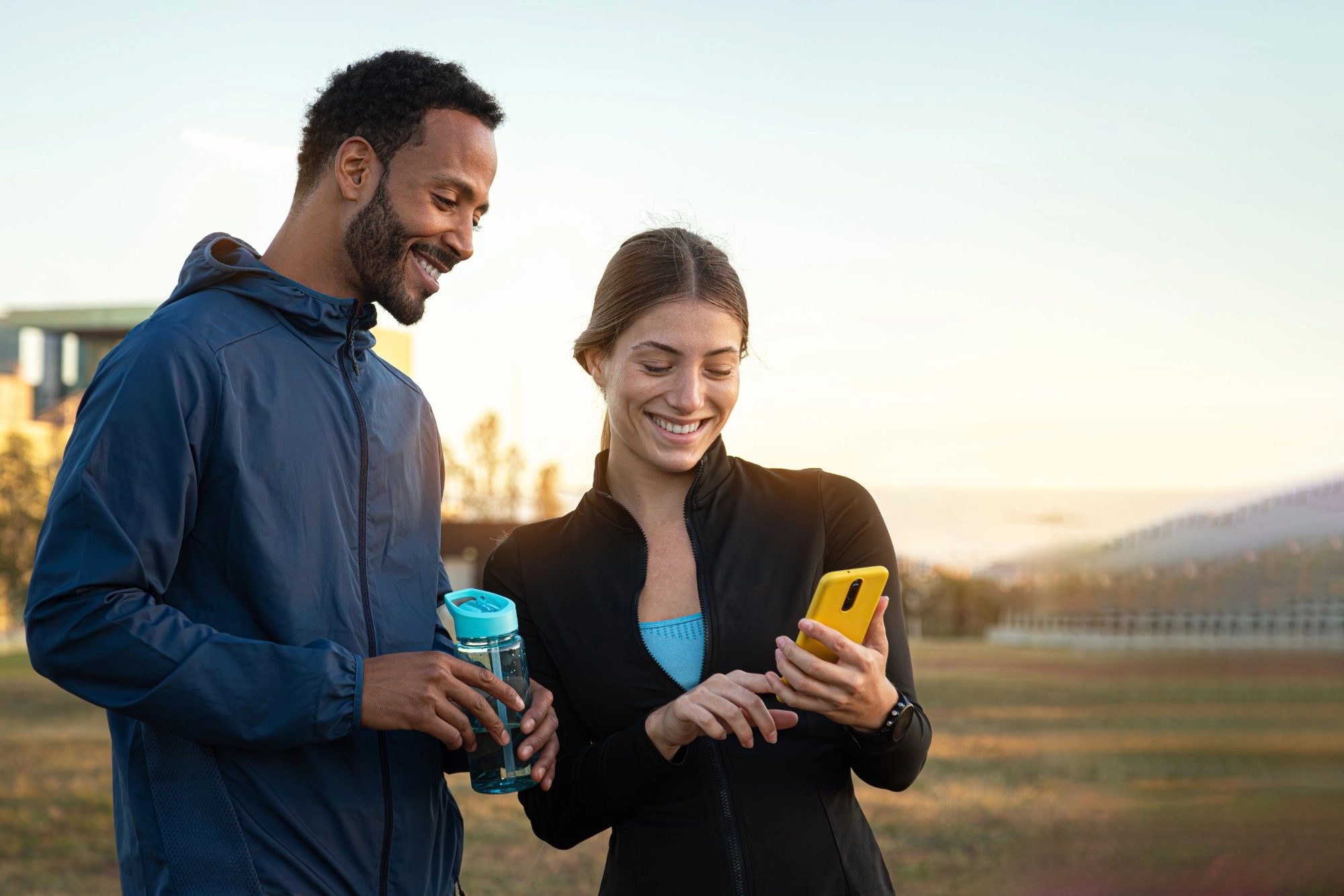 A smiling man and woman in running jackets looking at a yellow phone together outdoors