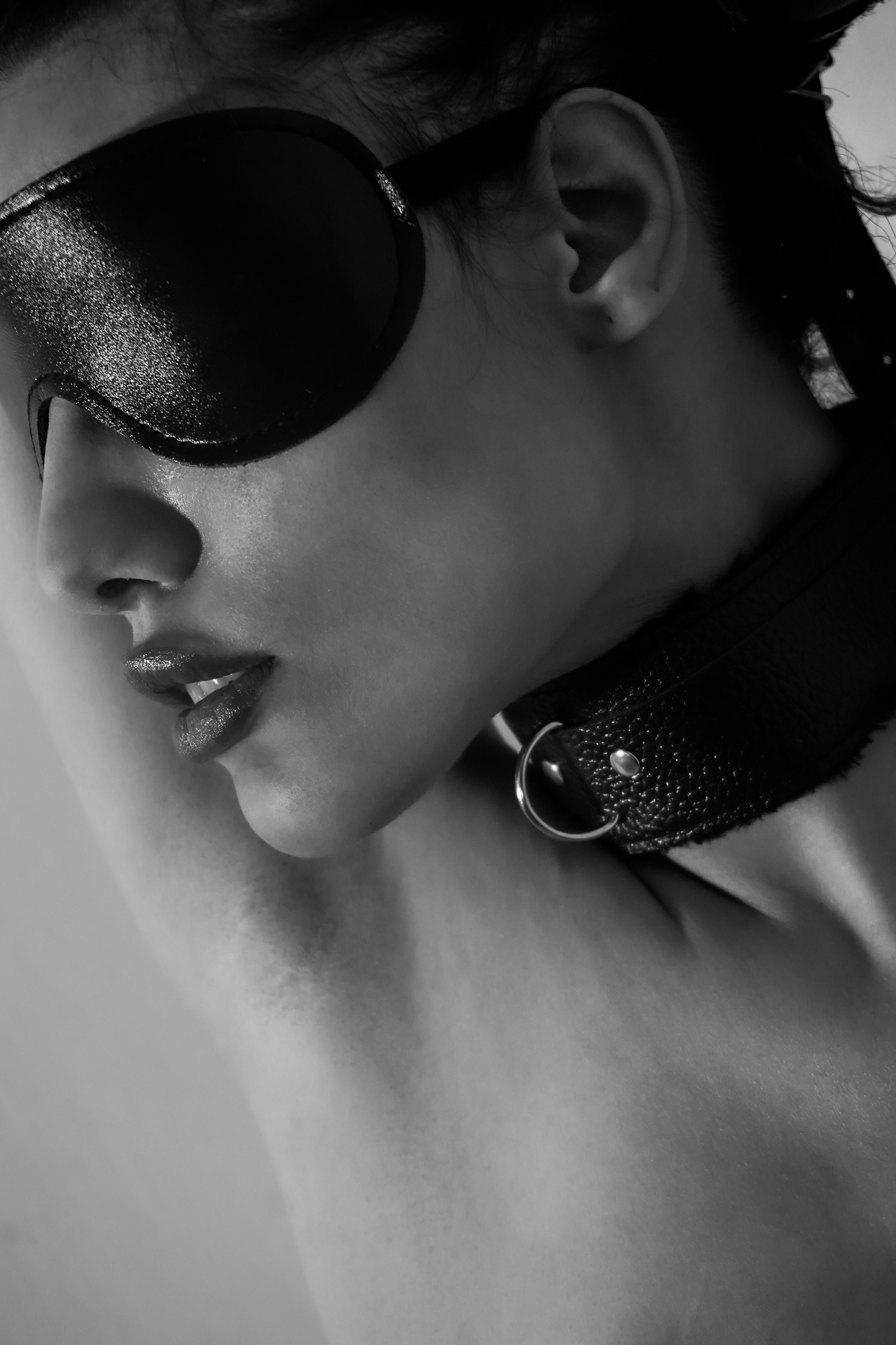 Black-and-white close-up profile portrait of a woman wearing a leather blindfold and a leather collar with an O-ring, bare shoulder visible