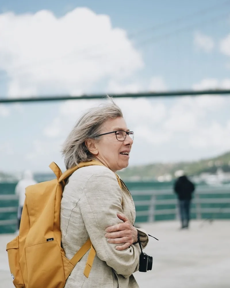 A woman with a yellow backpack and a camera looking out over a sunny waterfront with a bridge behind her
