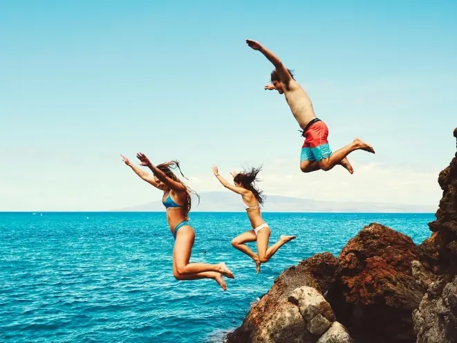 Three friends leaping off a rocky cliff into turquoise sea