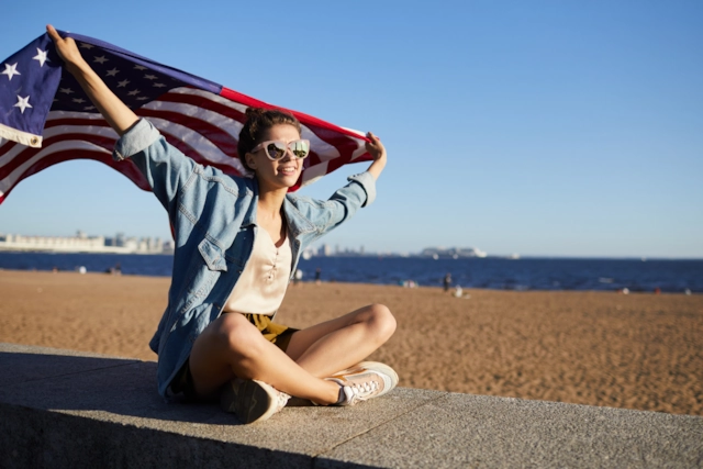 A woman in sunglasses sitting on a beach wall, holding the American flag up to the wind