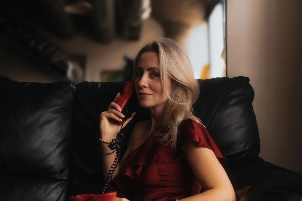 A blonde woman in a wine-red dress curled up on a black leather sofa, smiling as she talks on a vintage red telephone