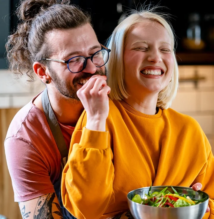 A man and a woman laughing together in a kitchen over a bowl of salad
