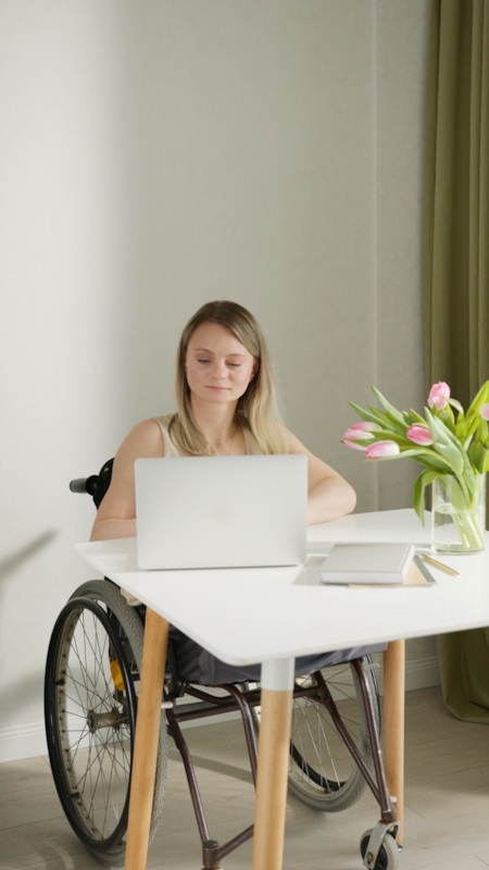 A woman in a wheelchair smiling while using a laptop at a white desk with a vase of pink tulips nearby