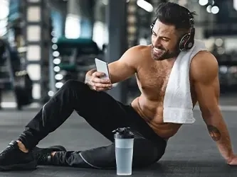 A smiling man with a towel and headphones checking his phone on the gym floor