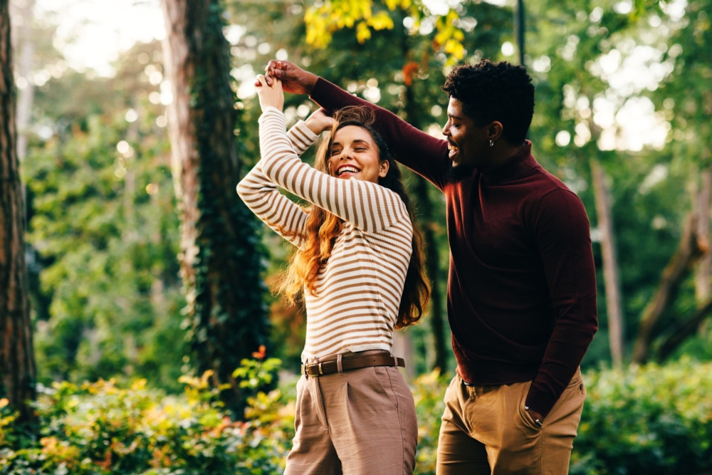 A man twirling a laughing woman as they dance in a leafy park
