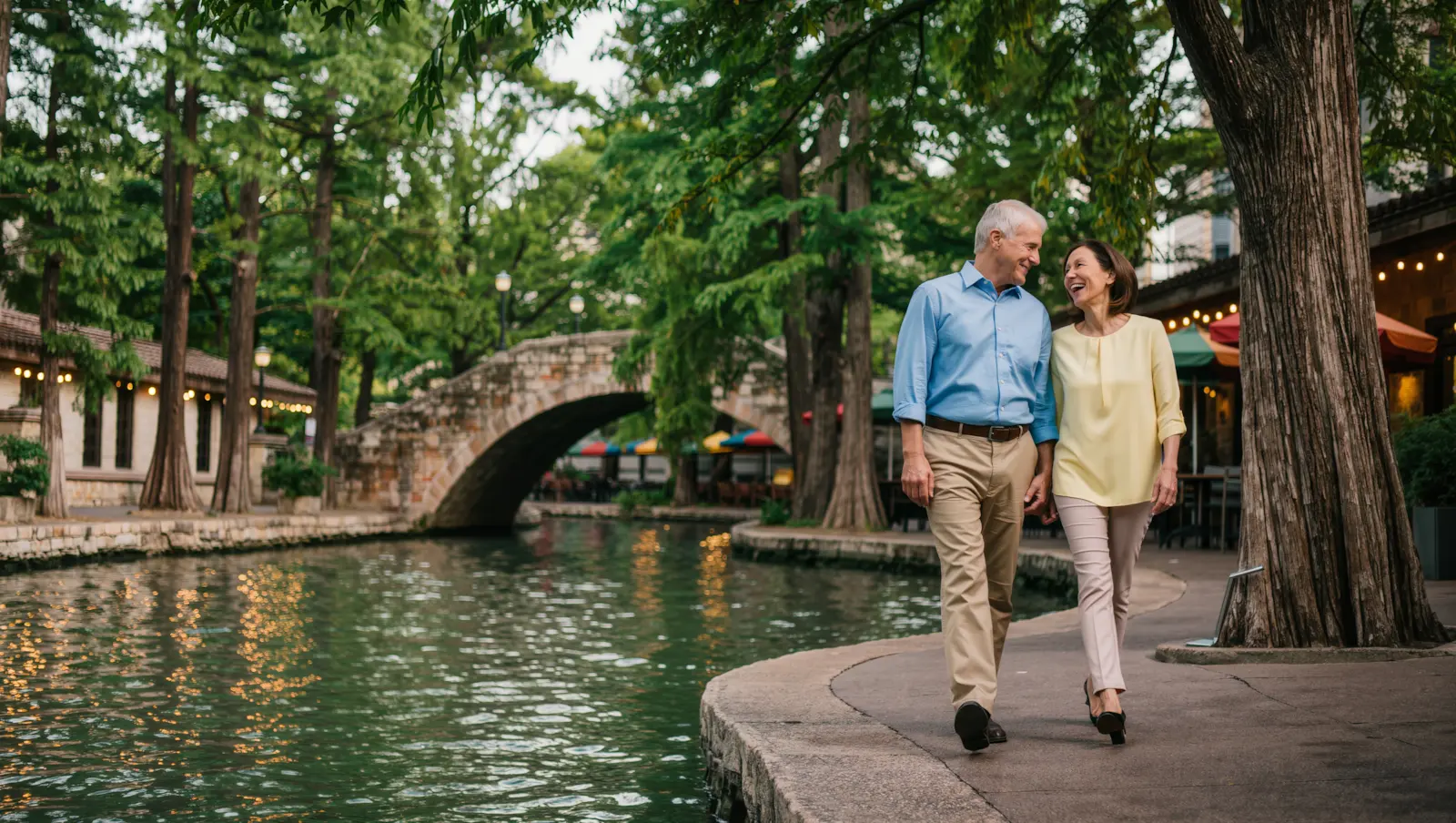 A couple in their late 50s laughing together as they walk beside the San Antonio River Walk in the evening, with a stone footbridge and string lights behind them