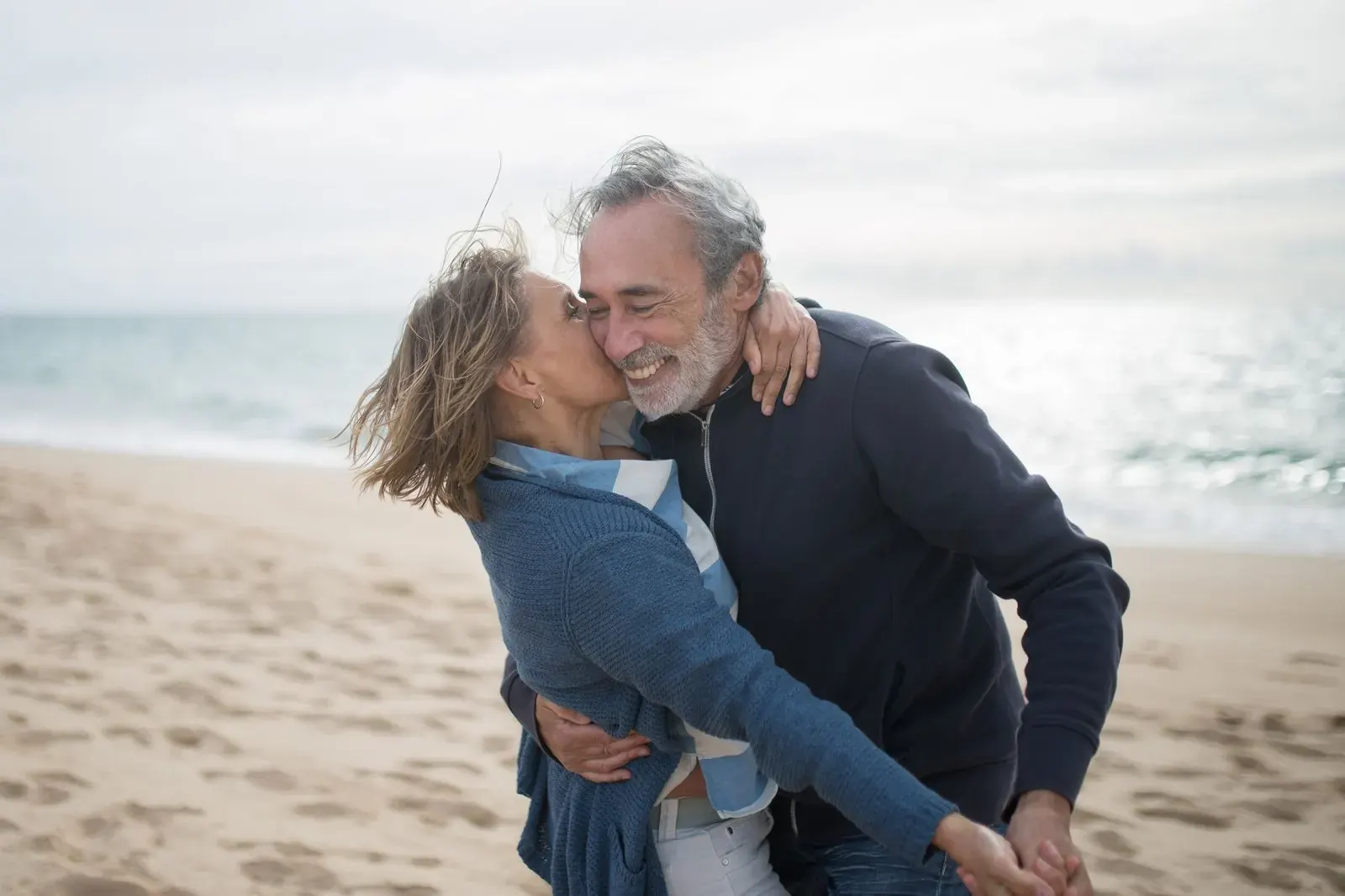 A woman kissing a laughing man on the cheek on a breezy beach