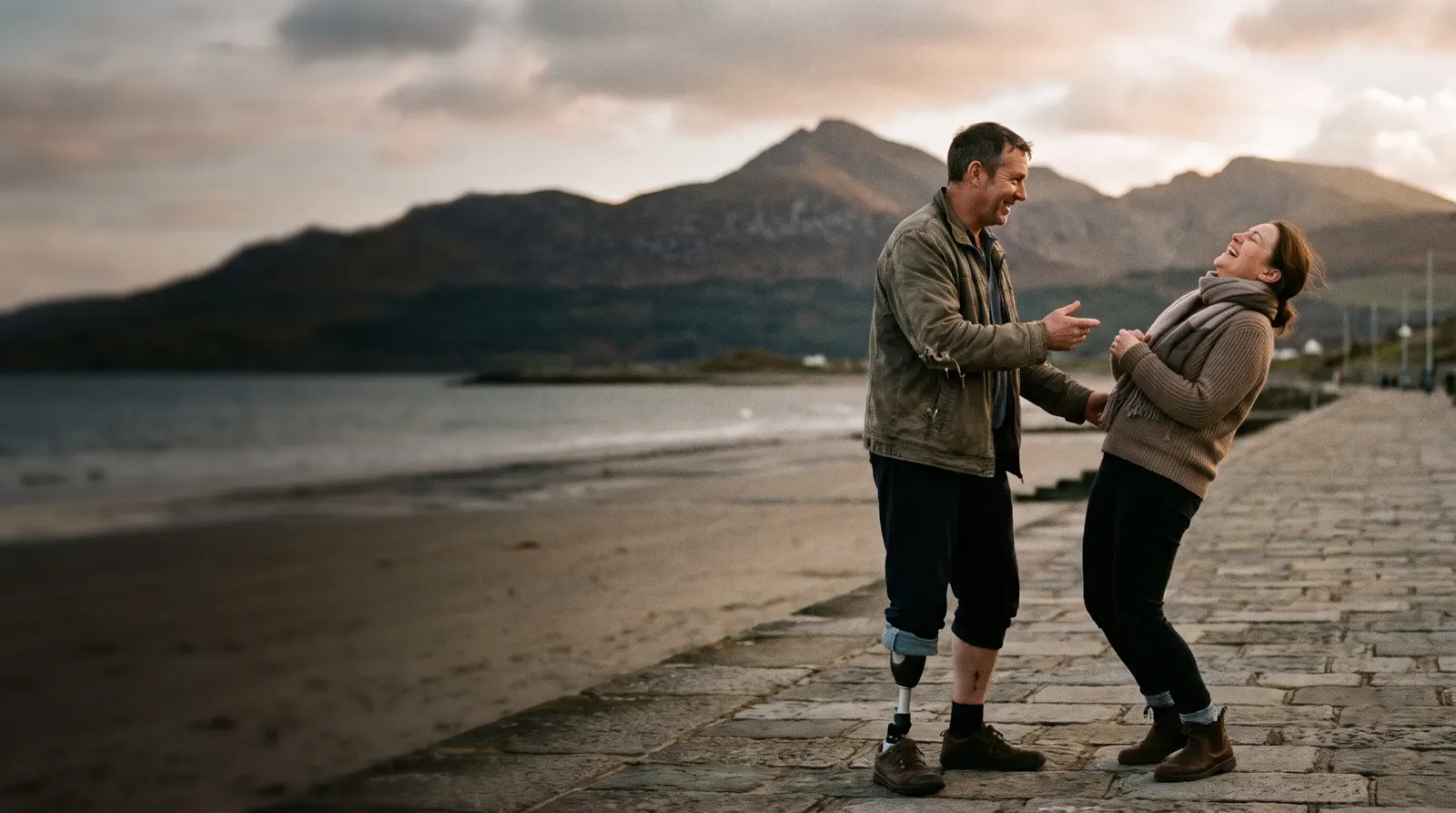 A couple laughing on the promenade at Newcastle, County Down, with the Mournes behind; the man has a prosthetic leg