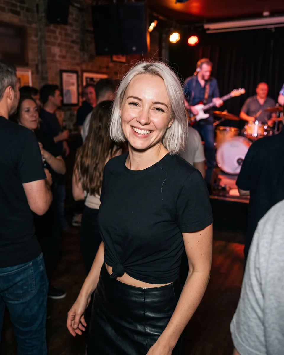 A woman with short platinum hair, a knotted black T-shirt and a leather skirt dances and grins in a packed Camden music bar, a blues band playing behind her (AI-generated image)