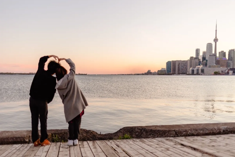 Couple making a heart with their hands on a pier, with the Toronto skyline and CN Tower at sunset