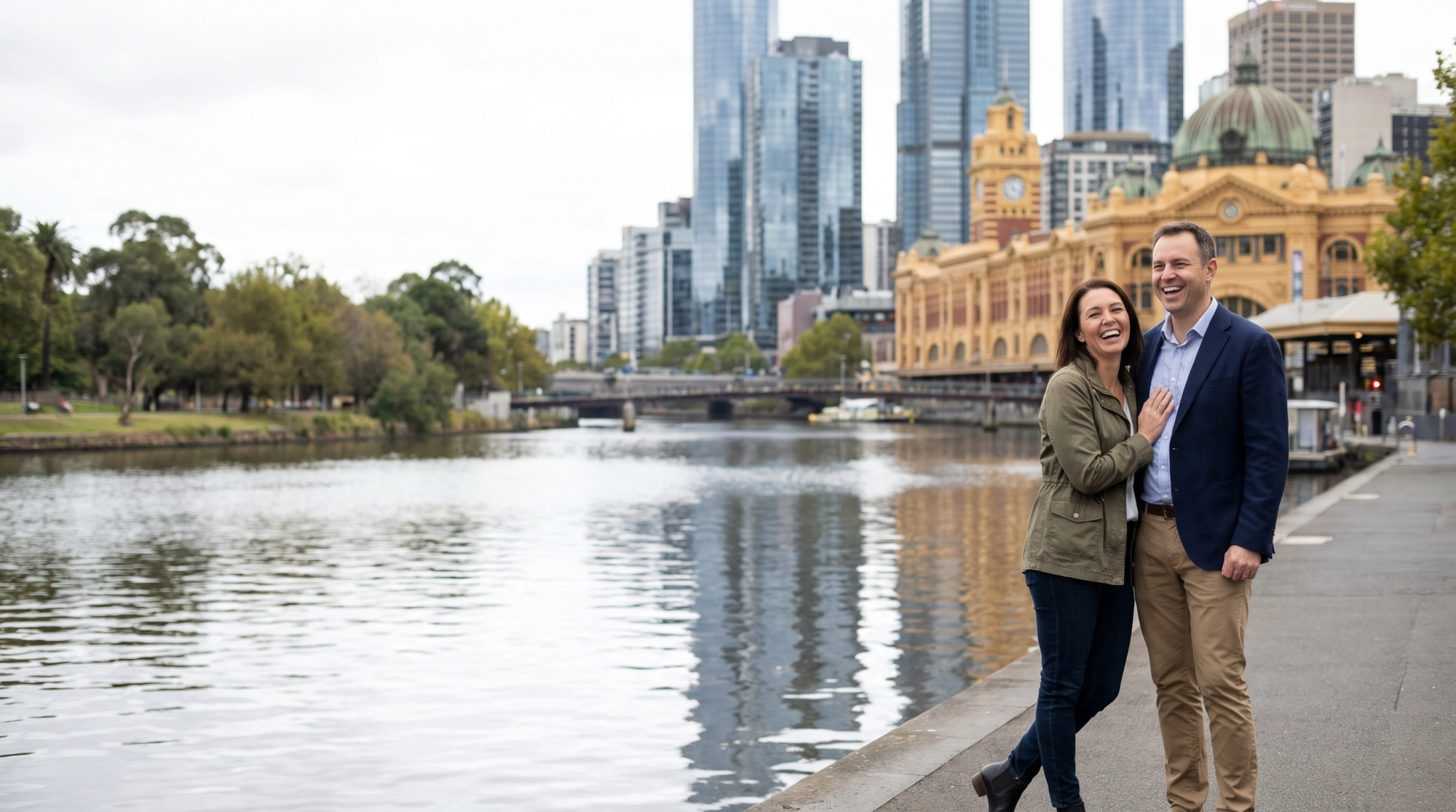 A happy couple laughing by the Yarra River with the Melbourne skyline and Flinders Street Station behind them