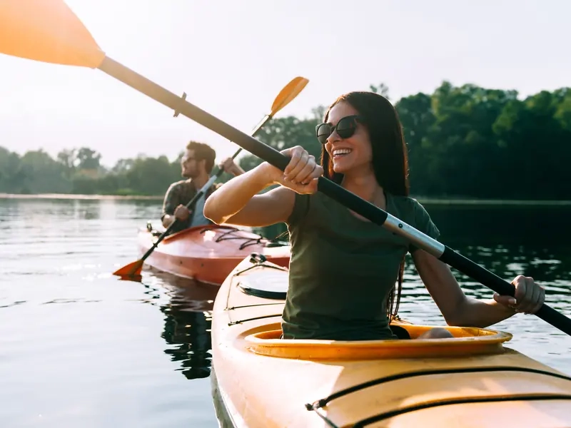 A smiling woman paddling a yellow kayak on a calm lake with a man kayaking behind her