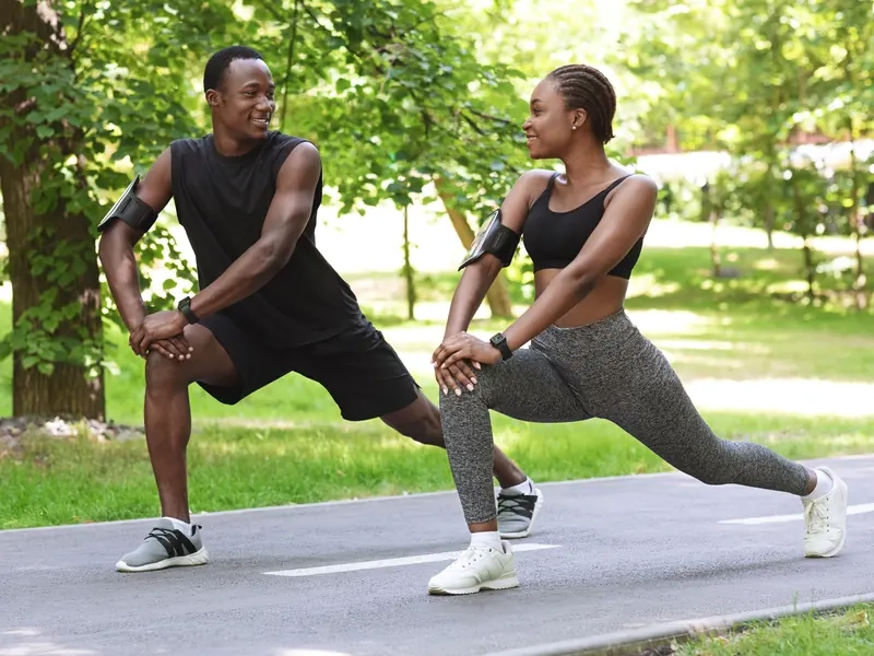 A smiling man and woman doing lunge stretches side by side on a sunny park path