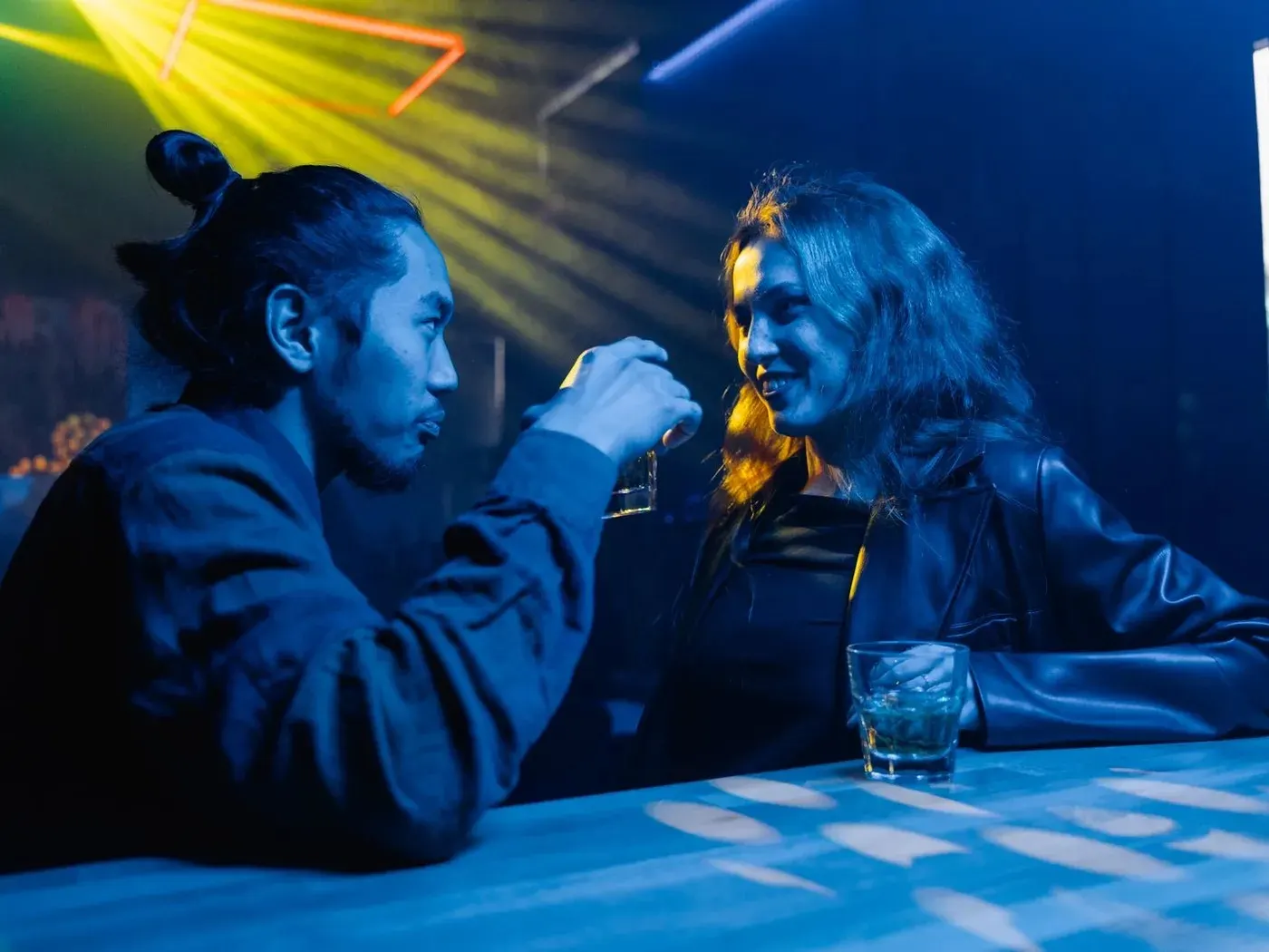 Two people flirting over drinks at a nightclub bar under blue and yellow club lights