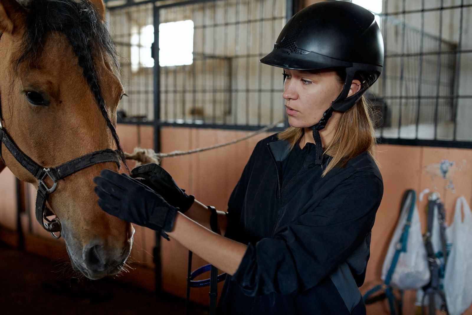 A rider in a black helmet and gloves adjusting a bridle on a horse in a stable block