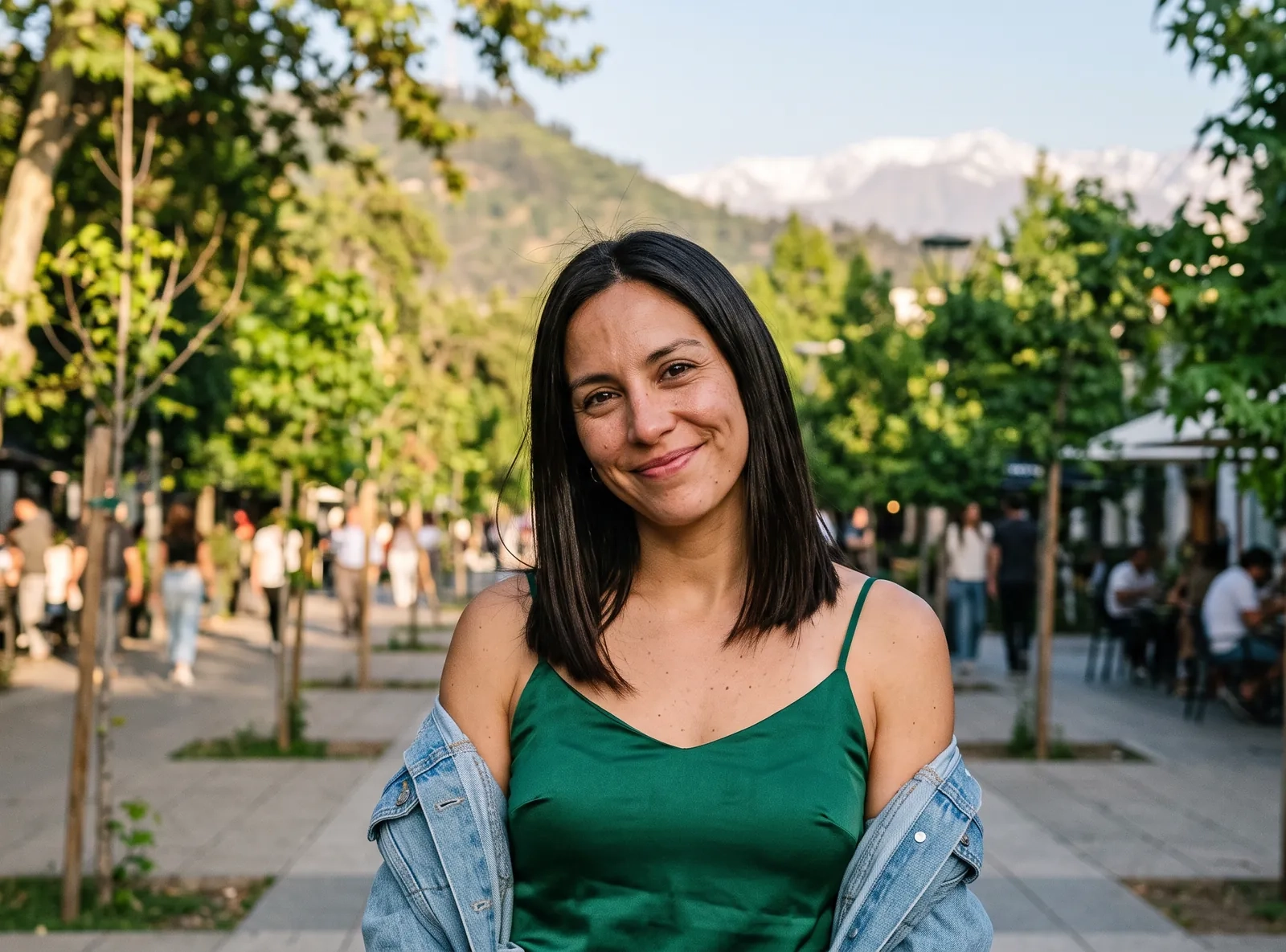 Una mujer de pelo liso castaño oscuro, con un top verde esmeralda y una chaqueta vaquera sobre los hombros, sonríe a la cámara en una calle peatonal arbolada de Santiago con la cordillera al fondo (imagen generada por IA)