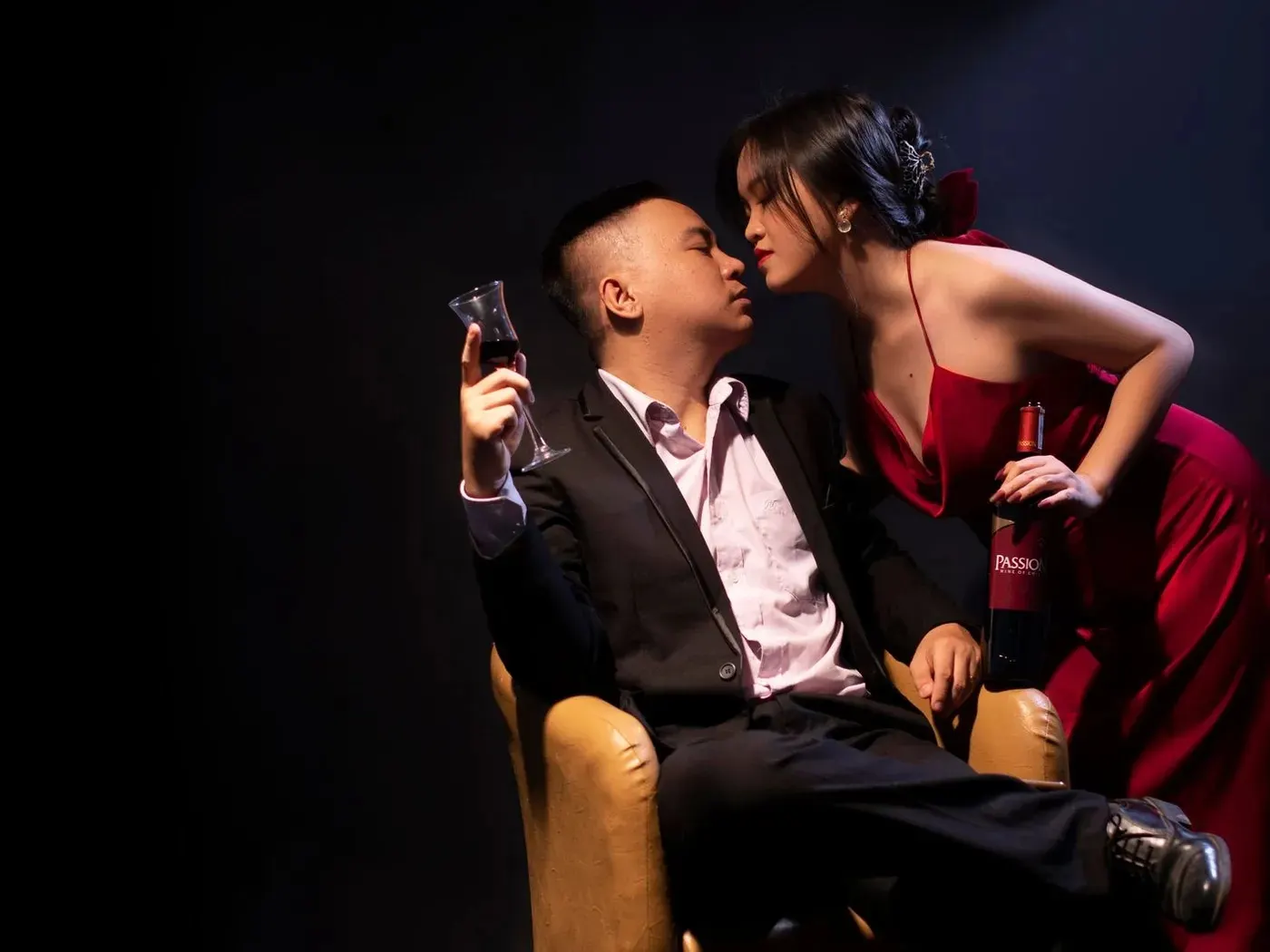 A woman in a red gown leaning in to whisper to a man in a dark suit sitting in an armchair with a glass of wine